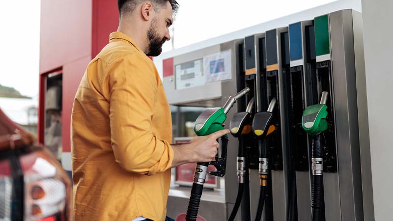 Pessoa abastece um veículo numa bomba de combustível, segurando uma mangueira verde, com várias bombas alinhadas e carros visíveis ao fundo.